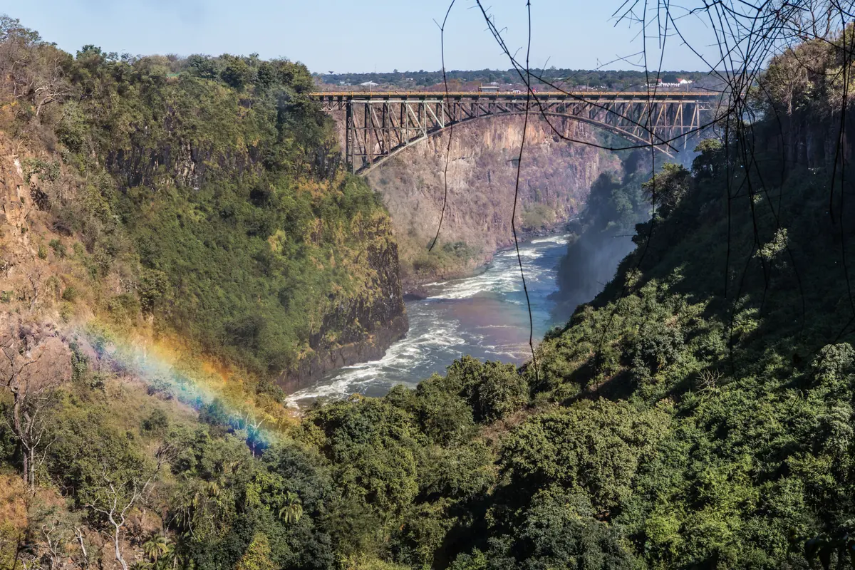 A steel arch bridge over a deep green gorge with a river below and a faint rainbow in the spray (the Victoria Falls Bridge, not the theatre)