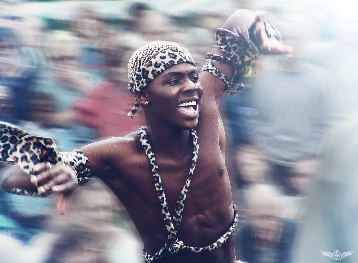A smiling dancer in a leopard-print headband and body harness with arms flung out, against a blurred crowd
