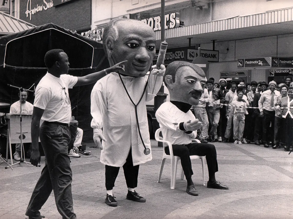 A black-and-white photograph of a 1980s street puppet show in Cape Town, with a puppeteer beside two large-headed puppets and a watching crowd (not Simunye)